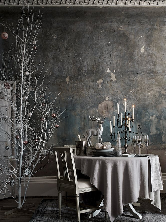 A festive dining setup with a white tablecloth, candles, a white reindeer figure, and Christmas ornaments on a dark, textured wall background.