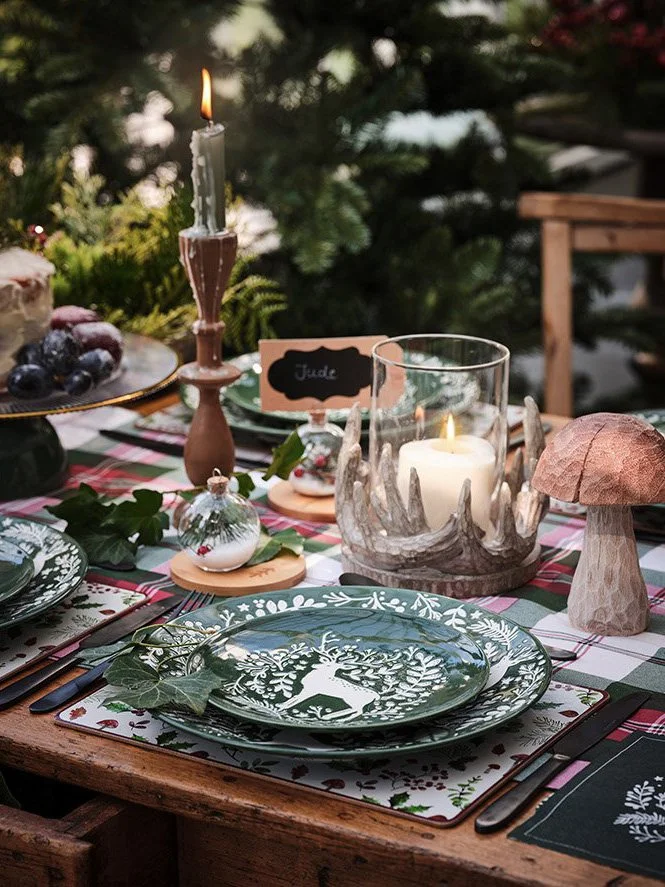 A festive holiday table setting with a green and white deer-themed plate, holiday ornaments, a small tree-shaped cake, a large candle in a decorative holder, and a candleholder with a lit candle, all on a plaid tablecloth with greenery and Christmas 