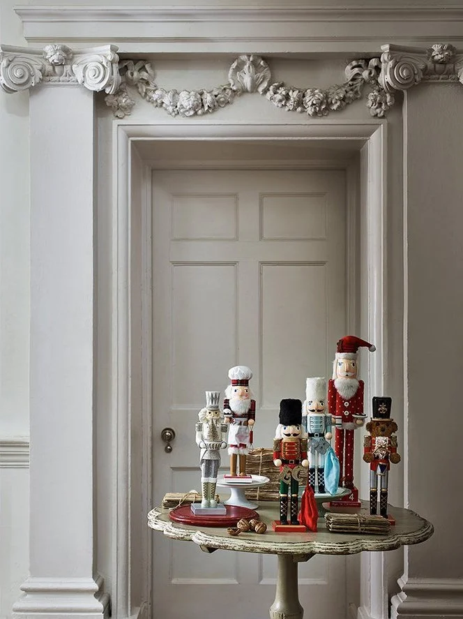 Christmas nutcracker figurines displayed on a vintage wooden table with a white door and ornate crown molding with floral garland in the background.