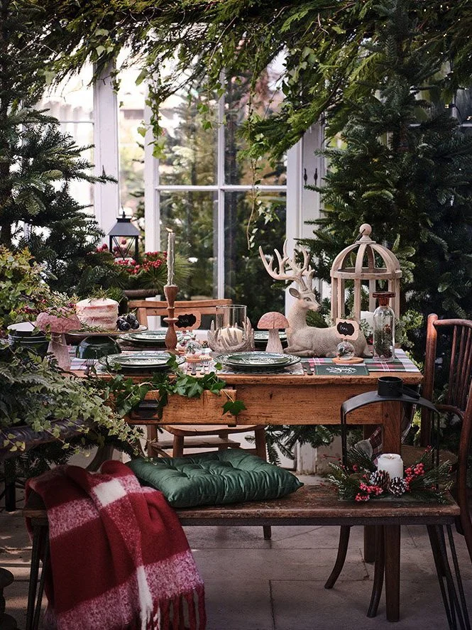 A cozy indoor Christmas setup with a wooden table decorated with Christmas-themed ornaments, a reindeer figure, a birdcage, and candles. The scene is surrounded by lush green foliage and trees, with a window in the background letting in natural light