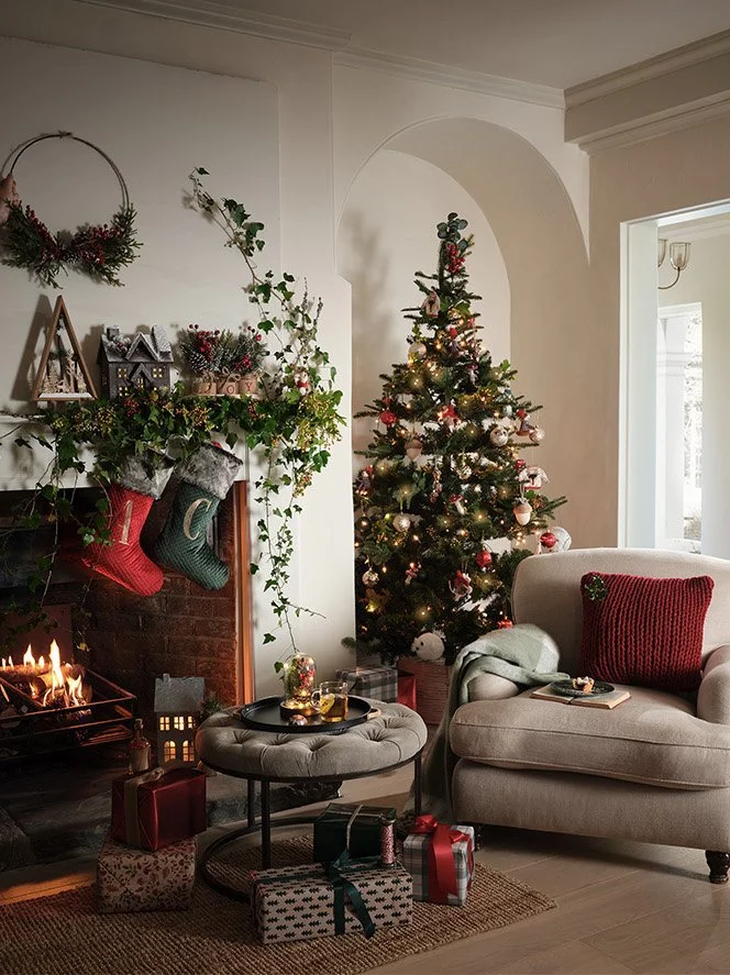 A cozy living room decorated for Christmas with a lit Christmas tree, a fireplace with stockings, wrapped presents, and comfy furniture.
