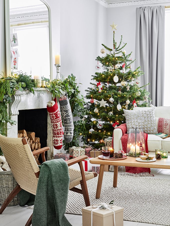 A decorated Christmas tree with ornaments, lights, and a star topper in a bright living room. A cozy fireplace with stockings hangs above, surrounded by greenery and candles. A white couch with cushions, a wooden coffee table with candles and snacks,