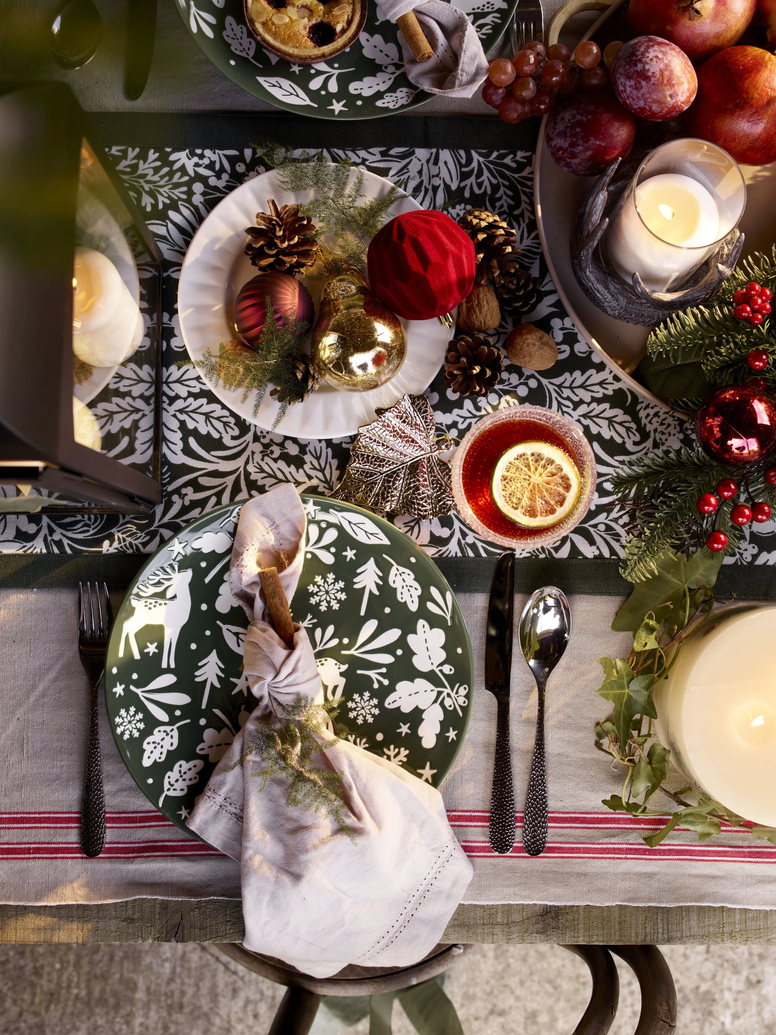 Festive holiday table setting with green and white patterned plates, Christmas ornaments, candles, and greenery.