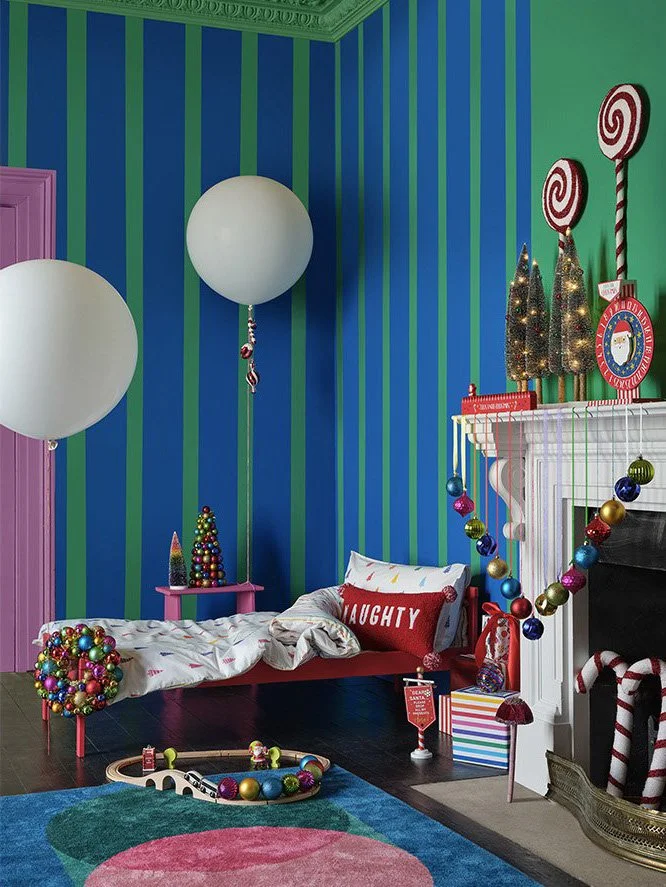 A festive Christmas-themed room with colorful striped walls, holiday decorations, and a bed with a pillow labeled 'Naughty'. There are Christmas trees, ornaments, and candy cane motifs, creating a playful holiday atmosphere.