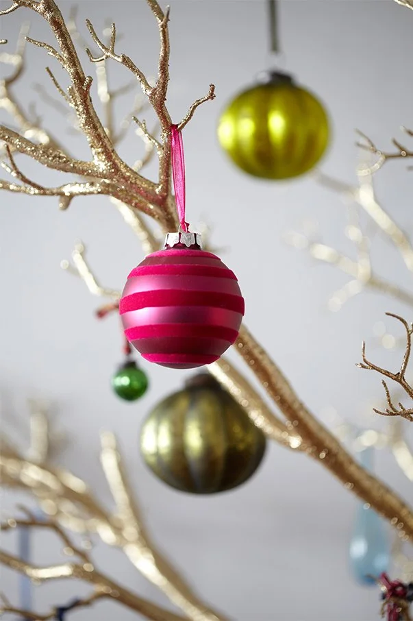 Pink and red striped Christmas ornament hanging on a gold-tinted, glittery tree branch, with additional green and gold ornaments in the background.