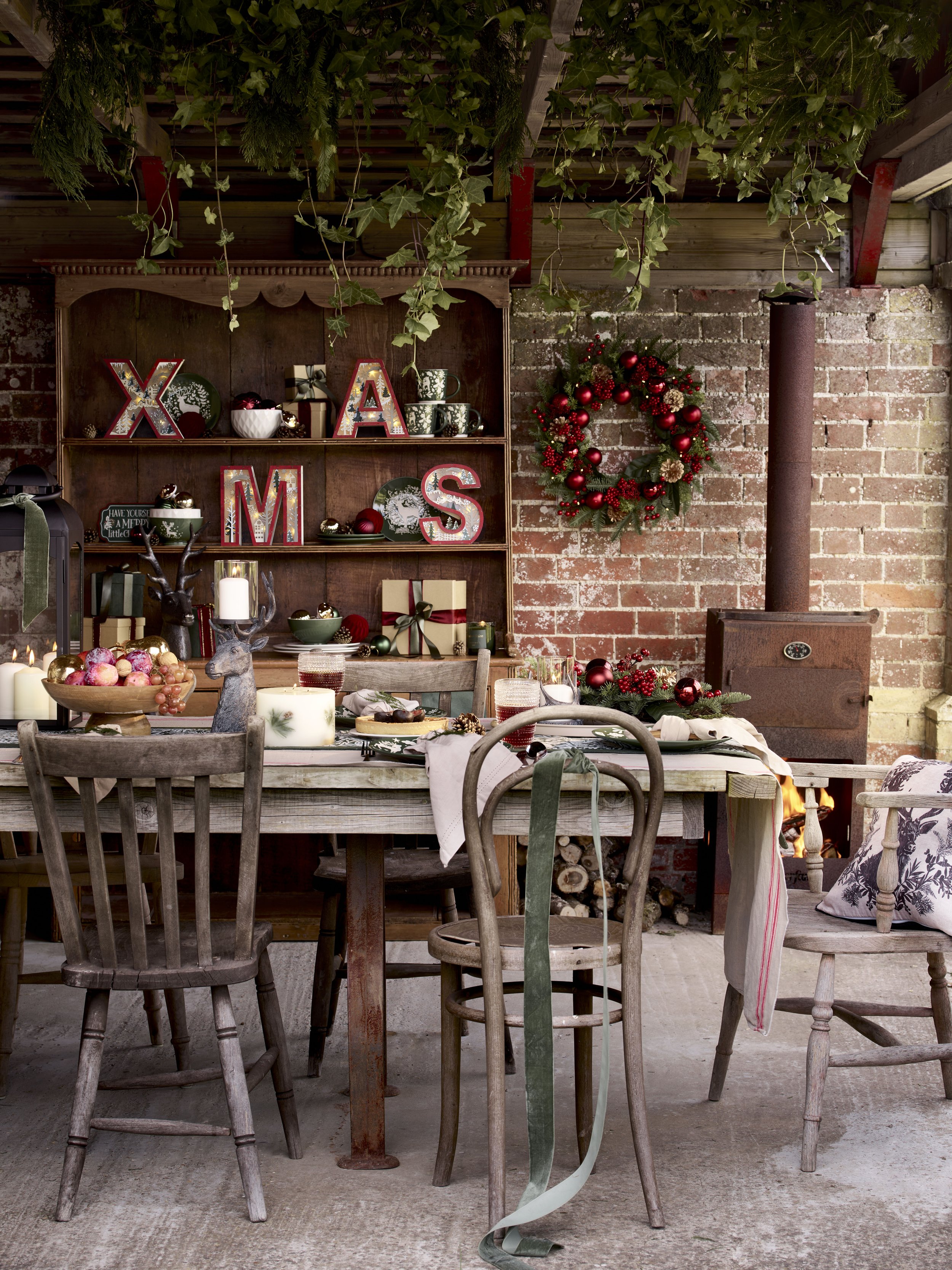 Rustic indoor Christmas scene with a wooden table, chairs, holiday decorations including a Christmas wreath, red and gold ornaments, wrapped gifts, and a brick wall with a wood and brick stove.