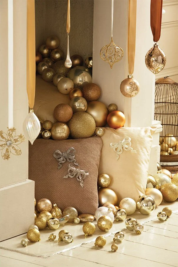 Decorative Christmas ornaments in gold, silver, and bronze colors are arranged around a cozy nook with pillows, creating a festive display.
