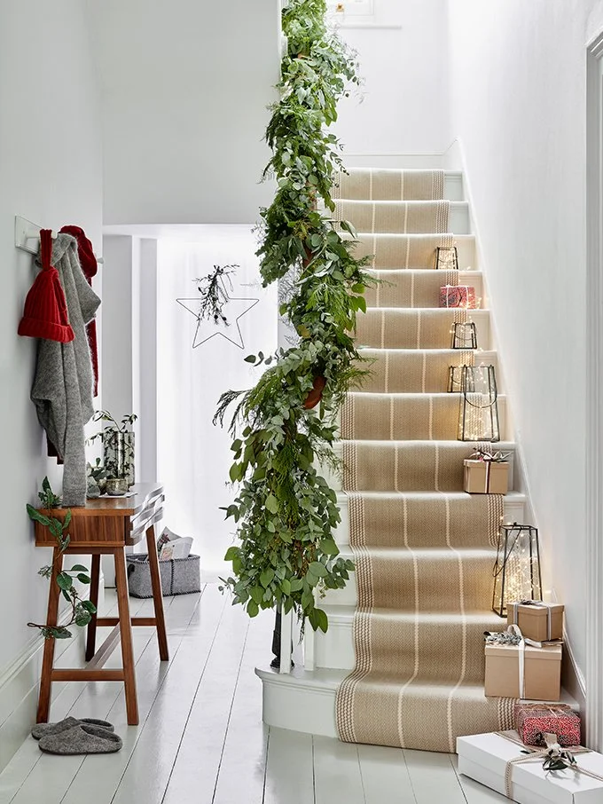 Interior of a home with a staircase decorated for Christmas, including wrapped gifts, lanterns, and a green garland with ornaments.