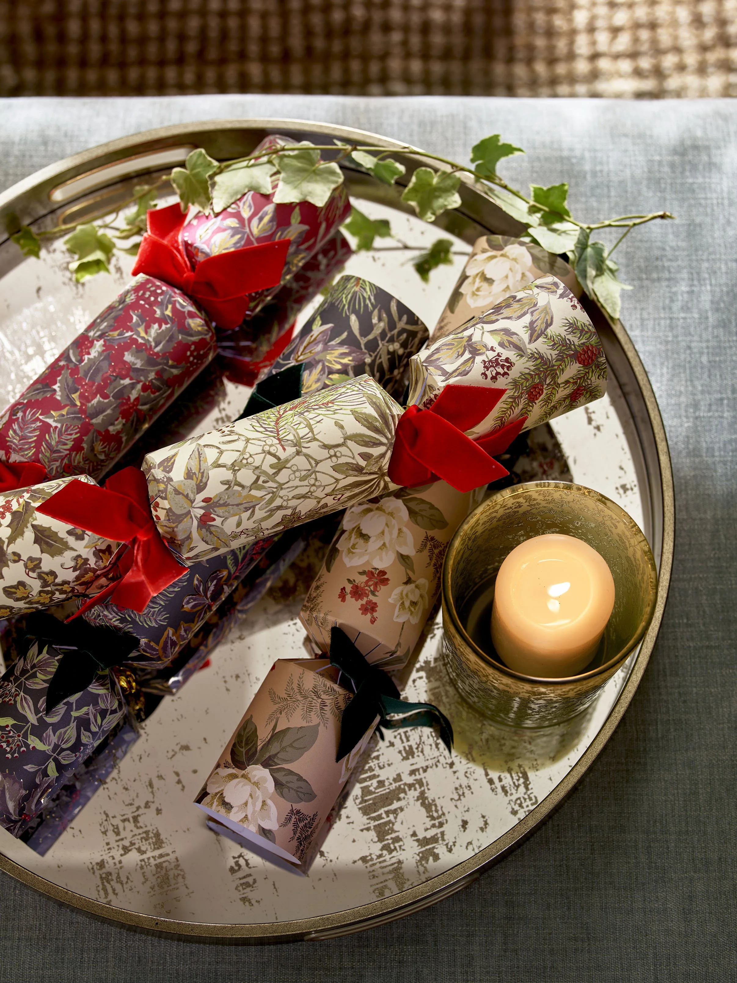 A tray holding wrapped Christmas gifts with floral and holiday patterns, tied with red and black ribbons, and a lit candle in a gold holder, on a table with a blue tablecloth and a decorative ivy plant.