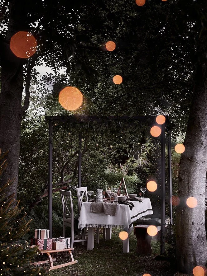 Outdoor dining table set for a meal in a lush garden, decorated with a white tablecloth, candles, and chairs, with blurred string lights in the foreground.