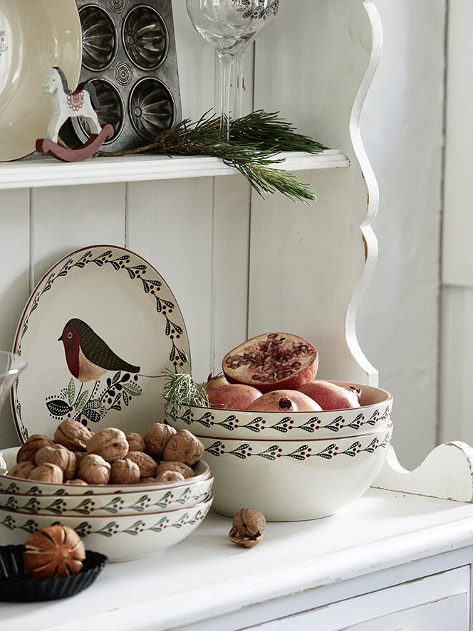 Ceramic bowls filled with hazelnuts and pomegranates on a white shelf with a decorative tray featuring a bird design, and a sprig of pine, set in a cozy, rustic kitchen environment.