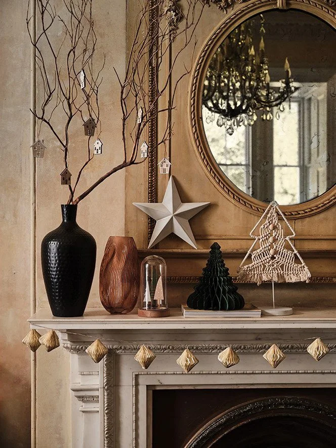 Decorative Christmas display on a fireplace mantel with vases, a star, a tree-shaped ornament, and hanging ornaments, with a mirror and chandelier reflected in the background.