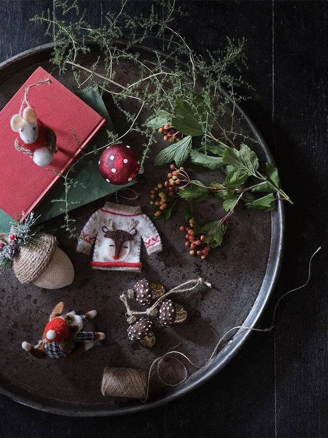 Holiday decorations on a round metal tray, including small animal plush ornaments, a red and white sweater with a reindeer, green leaves and berries, a red ornament, a red book, and craft supplies.