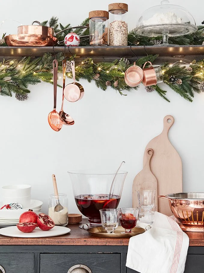 Kitchen countertop decorated for the holidays with a garland of pine branches, string lights, rose gold utensils hanging, glass jars with dry ingredients, a glass dome with baked goods, cutting boards, a bowl of red punch, pomegranate, glasses, and a