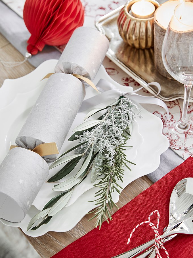 Festive holiday table setting with white plates, a sprig of faux frosted greenery, rolls wrapped in white paper with gold ribbons, red and white holiday decor, a silver tray, candles, a wine glass, and a red napkin with silverware tied with red and w