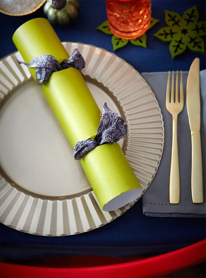 A festive table setting with a beige paper plate, a yellow rolled napkin secured with black patterned ribbons, a gold fork and knife on a gray napkin, a red glass, and a dark blue tablecloth.