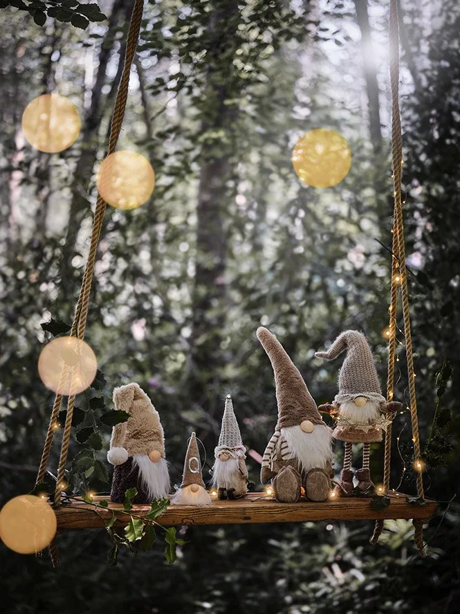 Decorative swing in a wooded outdoor setting with four plush gnome figurines dressed in festival attire, illuminated string lights, and blurred warm background lights.