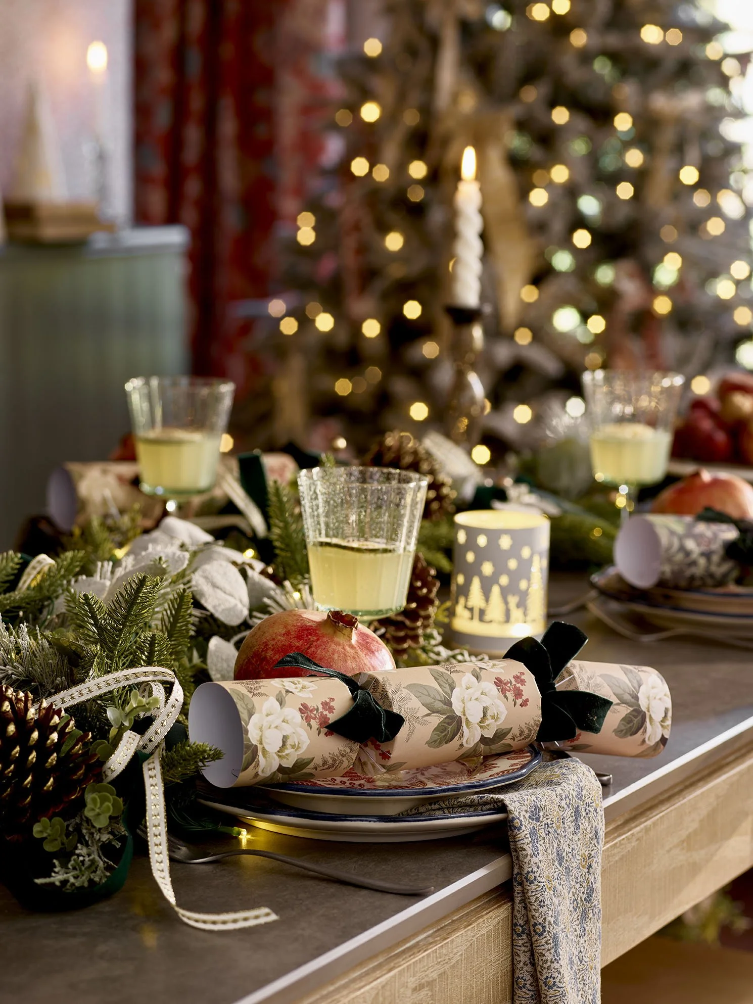 A festive holiday table setting with holiday decorations, wrapped gifts, glasses of white wine, and a Christmas tree in the background with blurred lights.