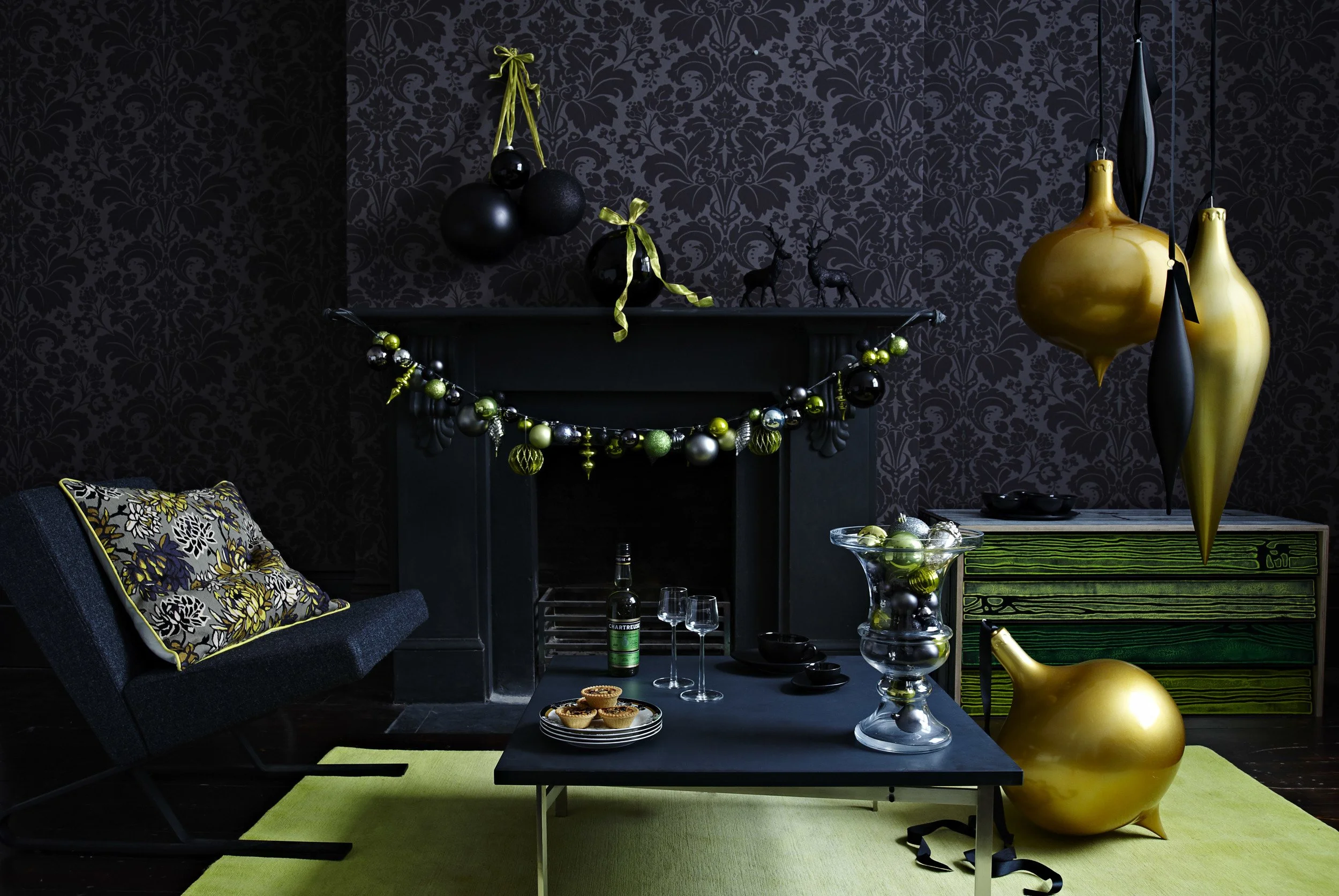 A living room decorated with black and gold holiday ornaments, including a pack of black balloons, a garland of black and green baubles, and oversized golden and black hanging ornaments. The fireplace mantel is decorated with small black reindeer fig