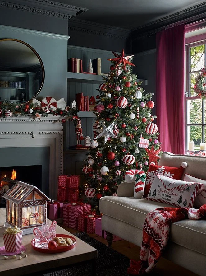 A decorated Christmas tree in a living room with red, white, and pink ornaments, wrapped gifts at the base, and holiday decorations on the mantle. There is a white couch with Christmas pillows and stockings, a lit fireplace, and a window with pink cu