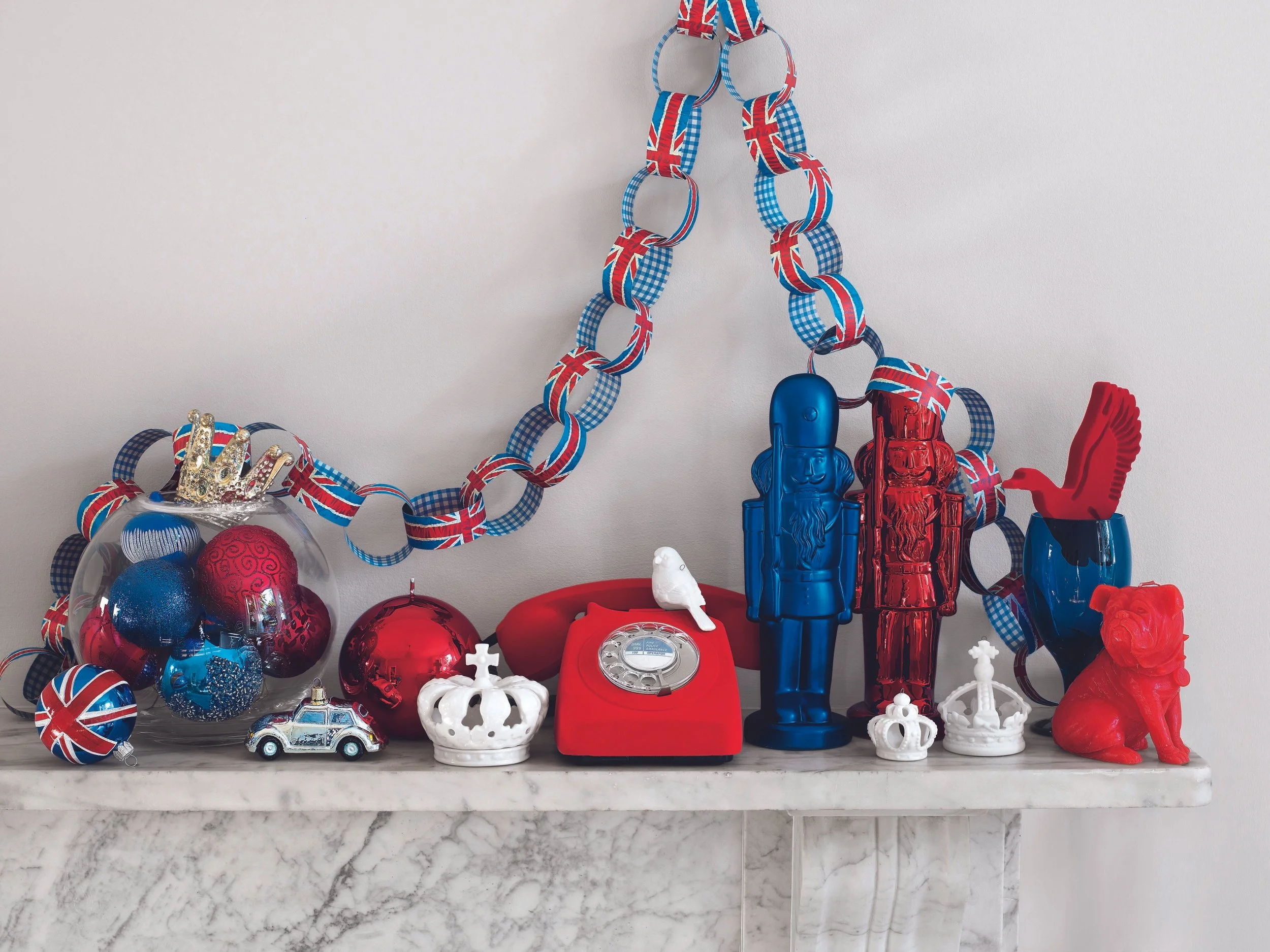Decorations with a Union Jack themed paper chain, Christmas ornaments, miniature crown and car, and British-themed figurines on a marble fireplace mantel.