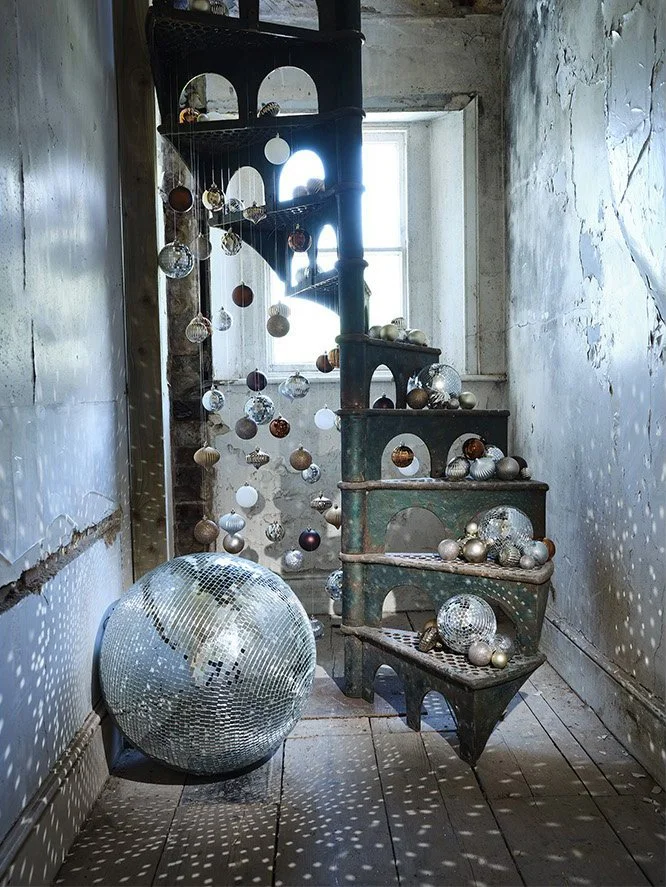 An old spiral staircase decorated with hanging ornaments and mirrored disco balls, with a large disco ball on the floor reflecting light onto the surrounding walls and ceiling in a rustic, worn room with peeling paint and a window in the background.