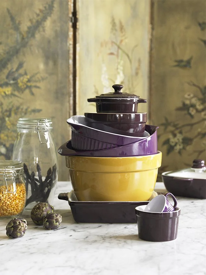 Stacked colorful ceramic baking dishes and bowls on a marble countertop, with jars of dried beans and artichokes nearby, set against a decorative background.