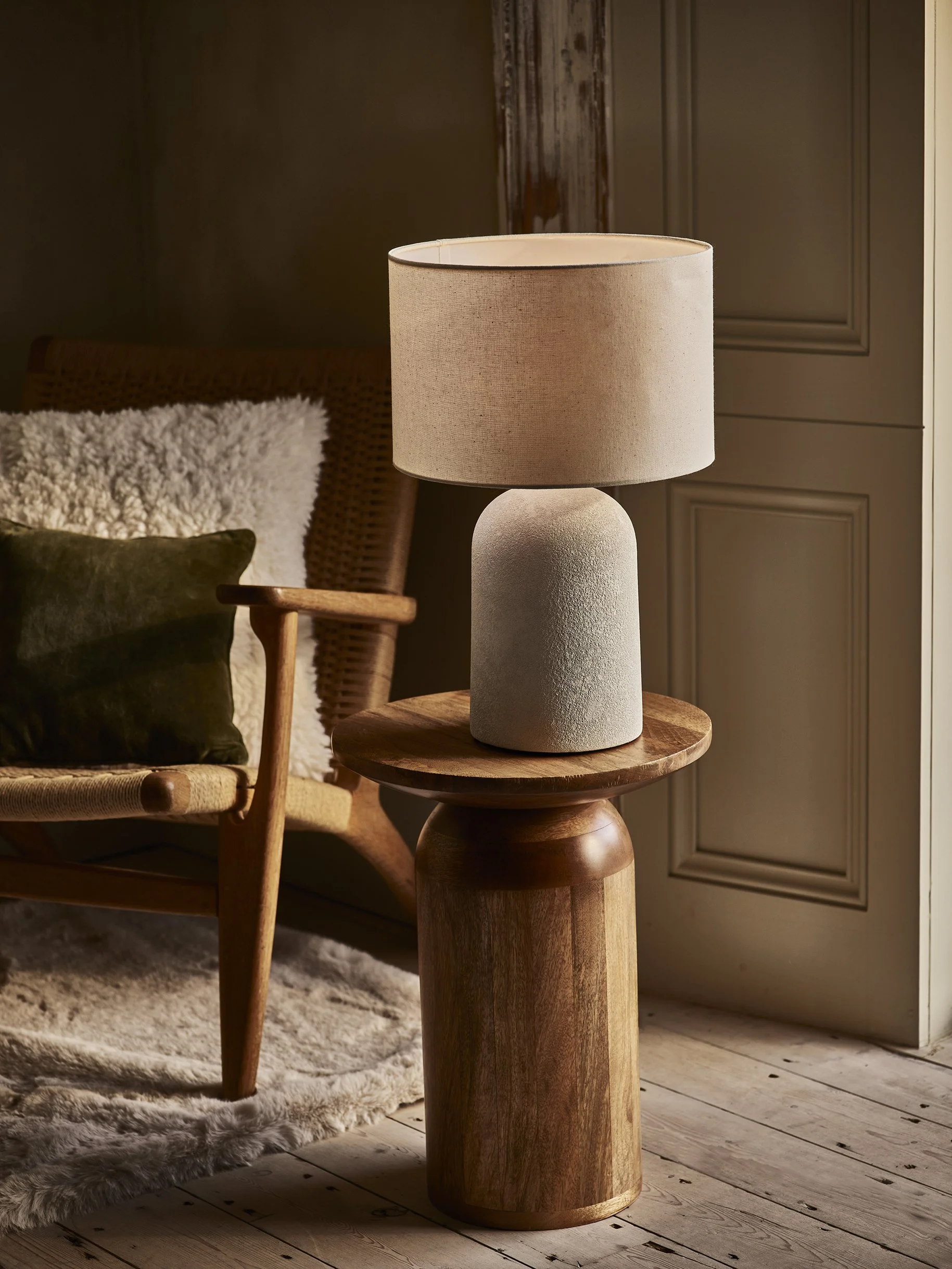 A beige table lamp with a cylindrical shade on a wooden side table in a cozy room with a bed and cushioned chair.