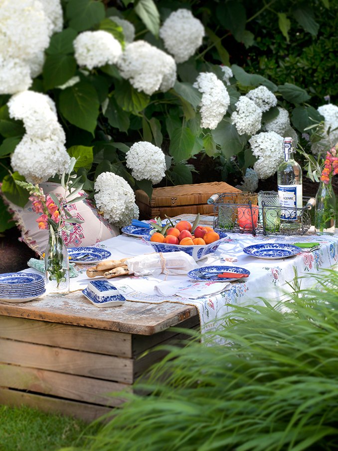 Outdoor table set for a meal with white hydrangeas in the background, featuring a white tablecloth, blue and white dishes, a bowl of peaches, and glassware, surrounded by lush greenery.