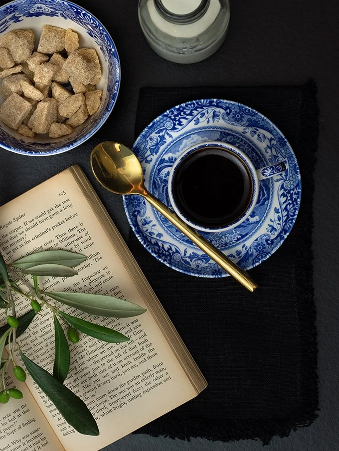 A table setting with a cup of black coffee on a blue floral patterned saucer, a gold spoon, an open book with green leaves on pages, a bowl of sugar cubes, and a bottle of milk or cream, all on a black placemat.