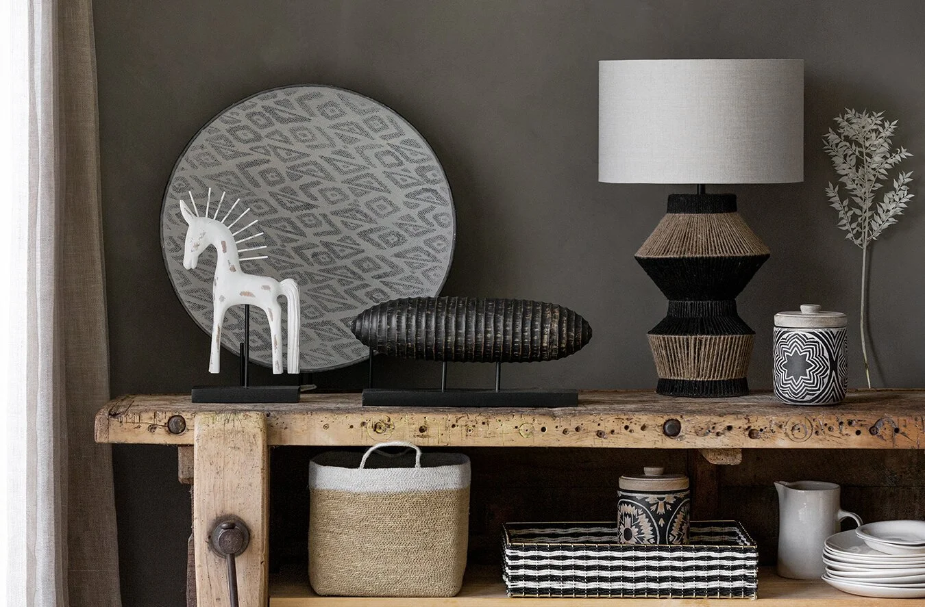 Decorative objects on a wooden table including a white ceramic giraffe, a round black and brown woven sculpture, a geometric ceramic lamp with a white shade, a ceramic jar with black and white star pattern, and a white leafy plant. There are addition