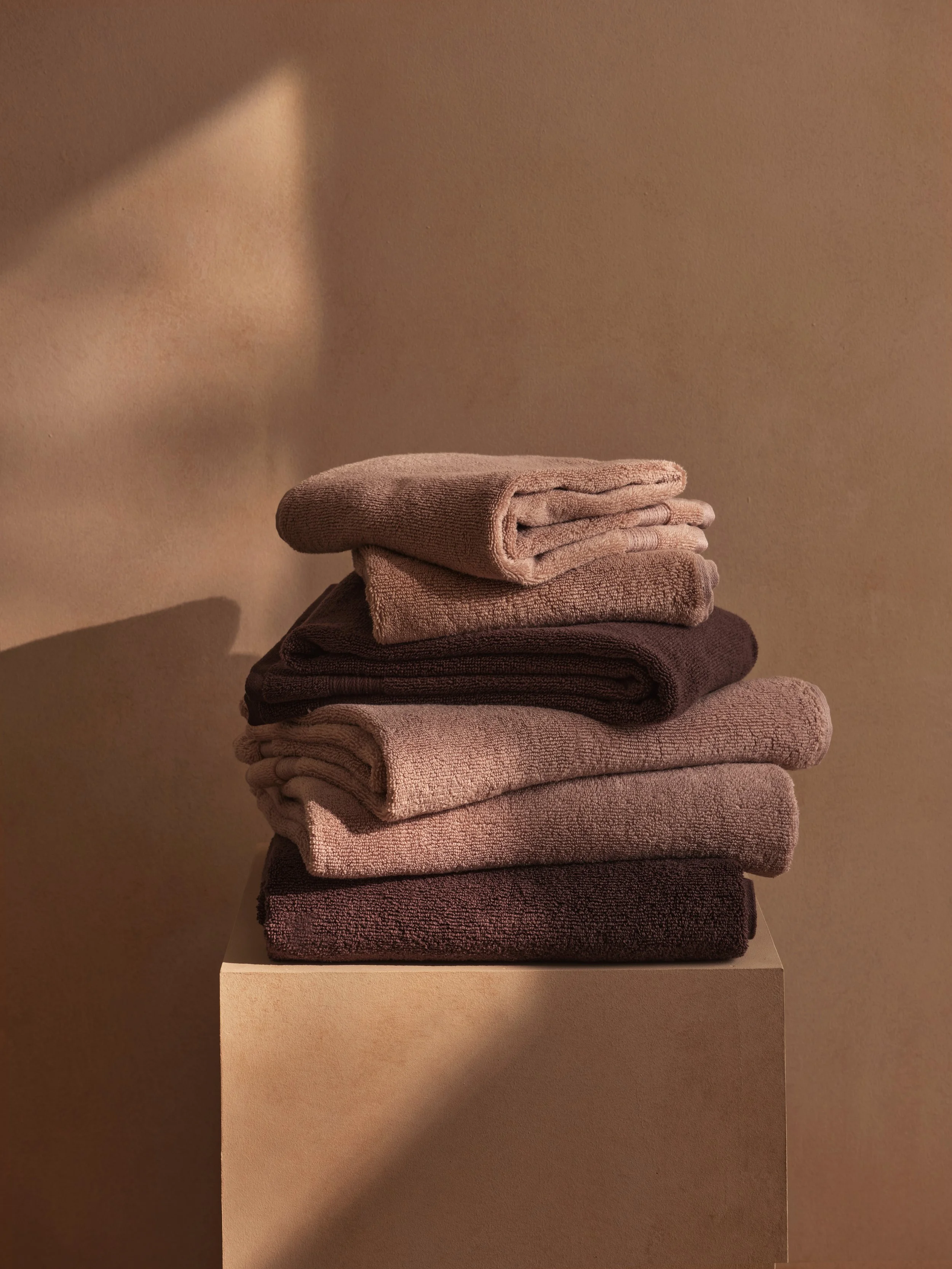 Stacked beige, brown, and gray folded towels on a neutral-colored pedestal with soft, warm lighting and shadows.