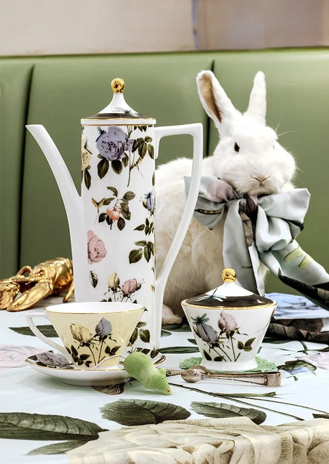 A white rabbit with a large bow tie sitting behind a floral tea set on a table, with a green cushioned bench in the background.