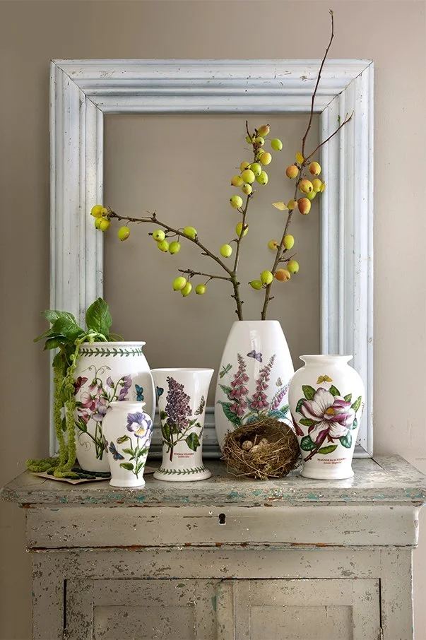 Six ceramic vases with floral and botanical designs on an old, distressed wooden surface, holding green and yellow berries, purple flowers, a green-leafed plant, and a bird's nest, all framed by a white picture frame resting behind them.