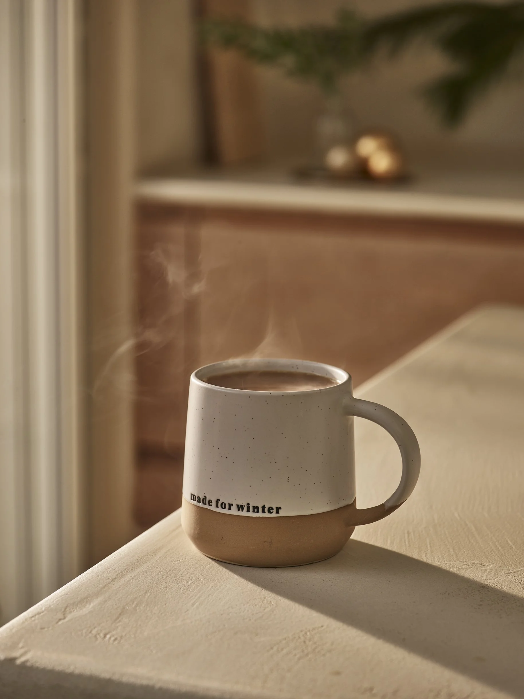 A ceramic mug filled with a hot beverage sitting on a table, with steam rising from it and sunlight casting a shadow. The mug has the text 'made for winter' written on it.