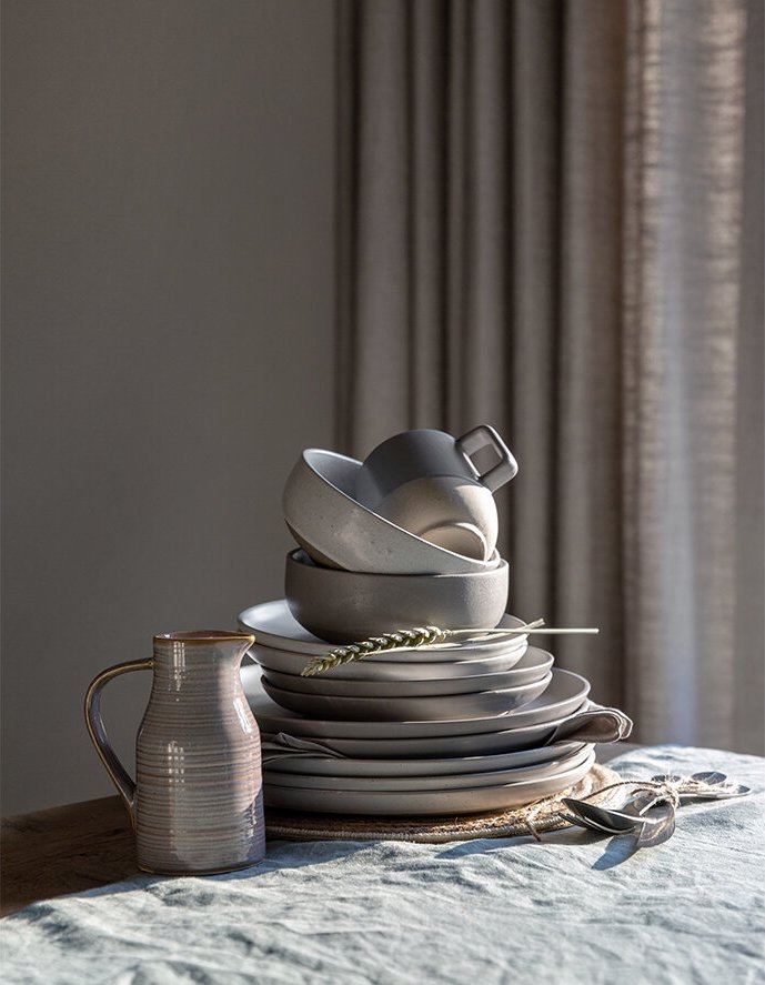 Stacked white and gray ceramic dishes and bowls with a ceramic pitcher on a table, sunlight coming through curtains in the background.