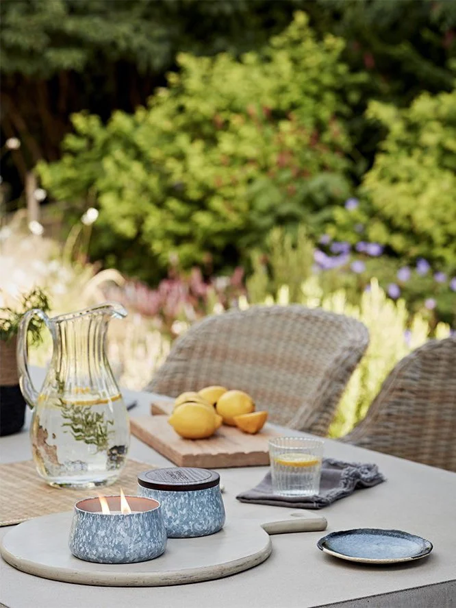Outdoor table set with candles, a glass of water with lemon, a wooden tray with lemons, a pitcher of water, and rattan chairs, with lush green trees and purple flowers in the background.