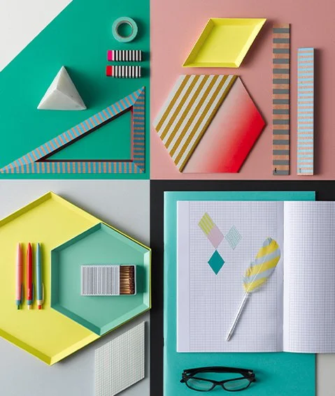 Flat lay of colorful stationery including geometric shapes, notebooks, pens, a ruler, an open grid notebook with paper shapes, and a pair of glasses.