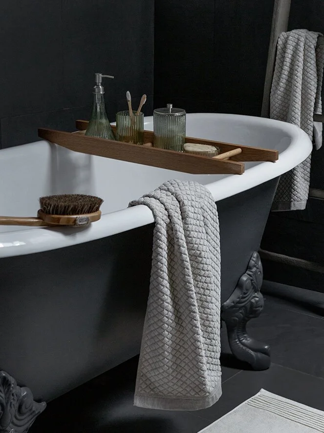 Bathroom with a black clawfoot tub and a gray textured towel hanging over the side, with a wooden tray holding soap, toothbrushes, and a glass container, on a dark tiled wall.