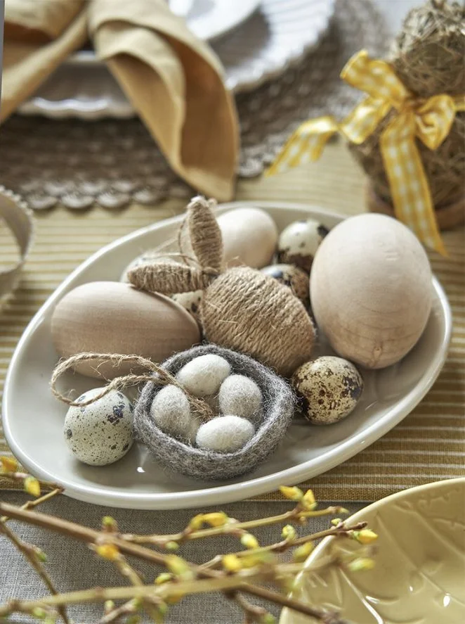 Decorative Easter eggs and natural elements on a white oval dish, surrounded by spring branches and tableware.