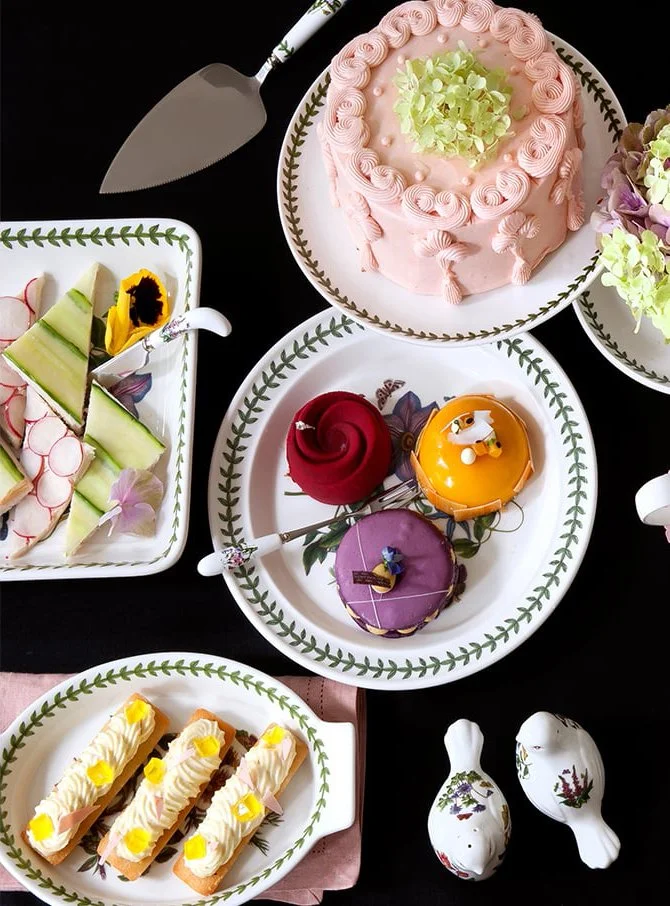A table with a pink frosted cake topped with green flowers, assorted colorful pastries, and fresh fruit slices on decorative plates and bowls.