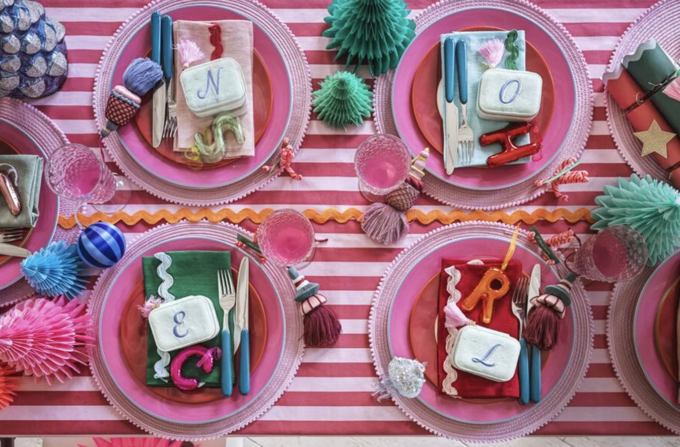 Party table setting with pink and red themed decorations, including pink placemats, decorative napkins, and pastel-colored spaghetti squash shaped tissue paper decorations. Each place setting has a small rectangular box with a letter and an attached 