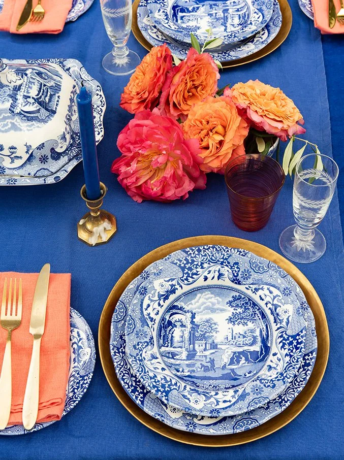 A table setting with blue and white patterned plates, orange napkins with gold utensils, a gold-rimmed charger plate, a blue candle in a gold holder, pink and orange peony flowers, a burgundy glass, and two clear glasses on a blue tablecloth.