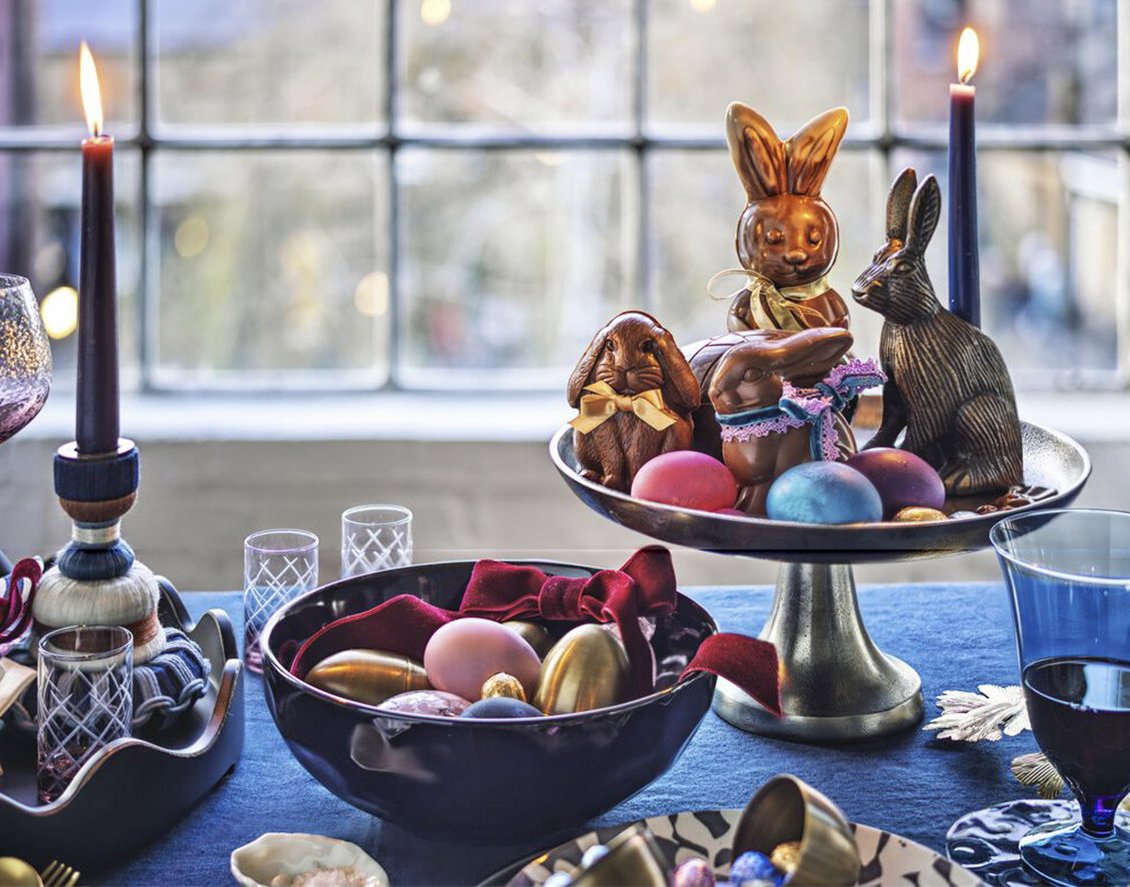 Decorative Easter table centerpiece with chocolate bunny and squirrel, colorful eggs, lit candles, and tableware, set in front of a window.