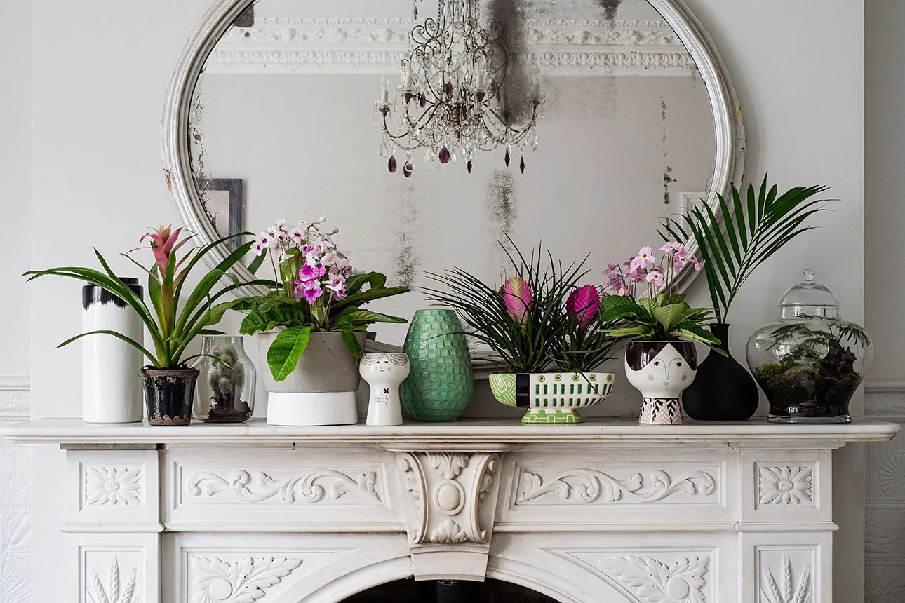 A white decorative fireplace mantel with various potted plants, vases, and decorative objects in front of a large round mirror and a chandelier reflected in the mirror.