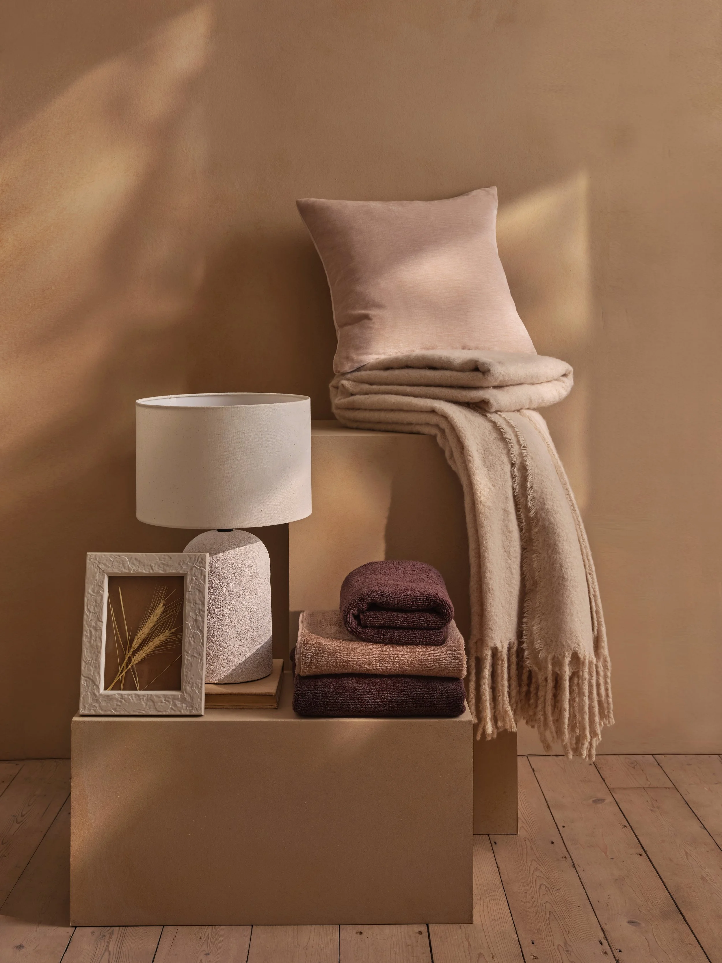 Decorative display with beige pillows and throw blankets stacked on a gold-colored pedestal, a white table lamp with textured base, a white picture frame with a gold plant photograph, and folded towels in shades of beige and dark red on a wooden floo
