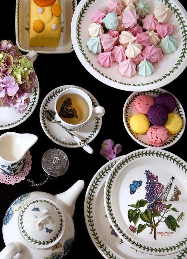 Assorted colorful macarons, meringues, tea, and floral tableware on a black table.