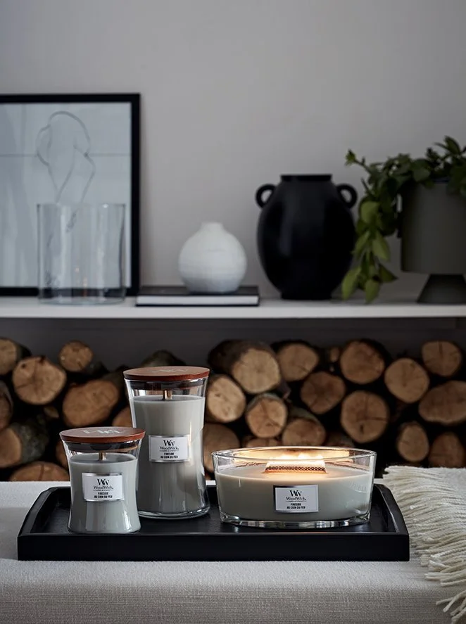A decorative living room shelf with glass vases, a black vase, and a potted plant, with logs stacked underneath; in front, there are three candles on a tray, two in glass containers and one in a glass dish.