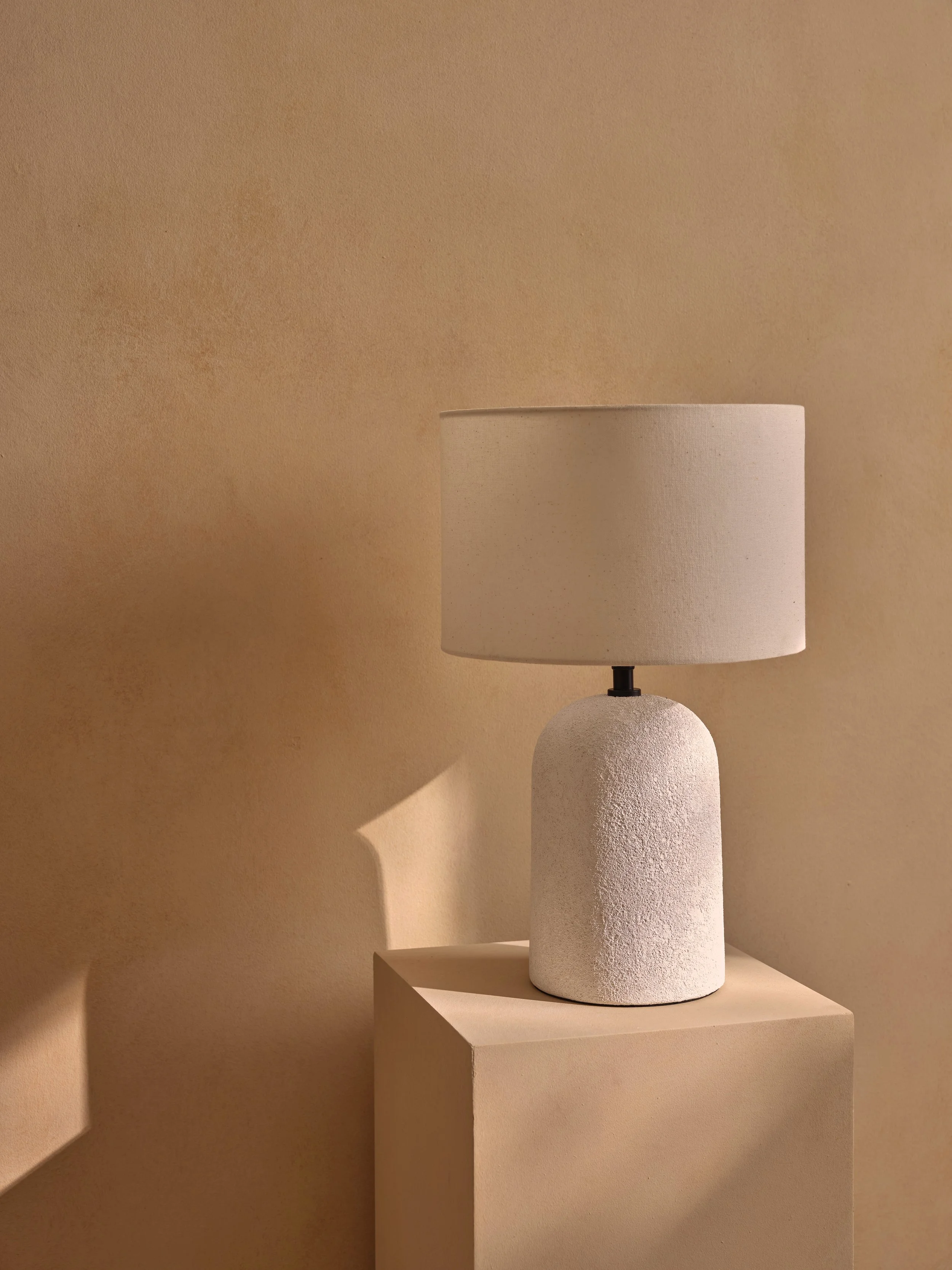 A white textured table lamp with a round base and a beige drum shade, placed on a beige pedestal against a beige wall.