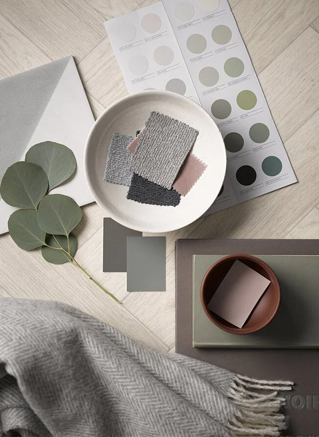 Flat lay arrangement of fabric swatches, color samples, and decor items including a white bowl with fabric pieces, a sprig of eucalyptus, color palette sheets, a gray book, and a small of brown bowl with a note, on a light wooden surface.